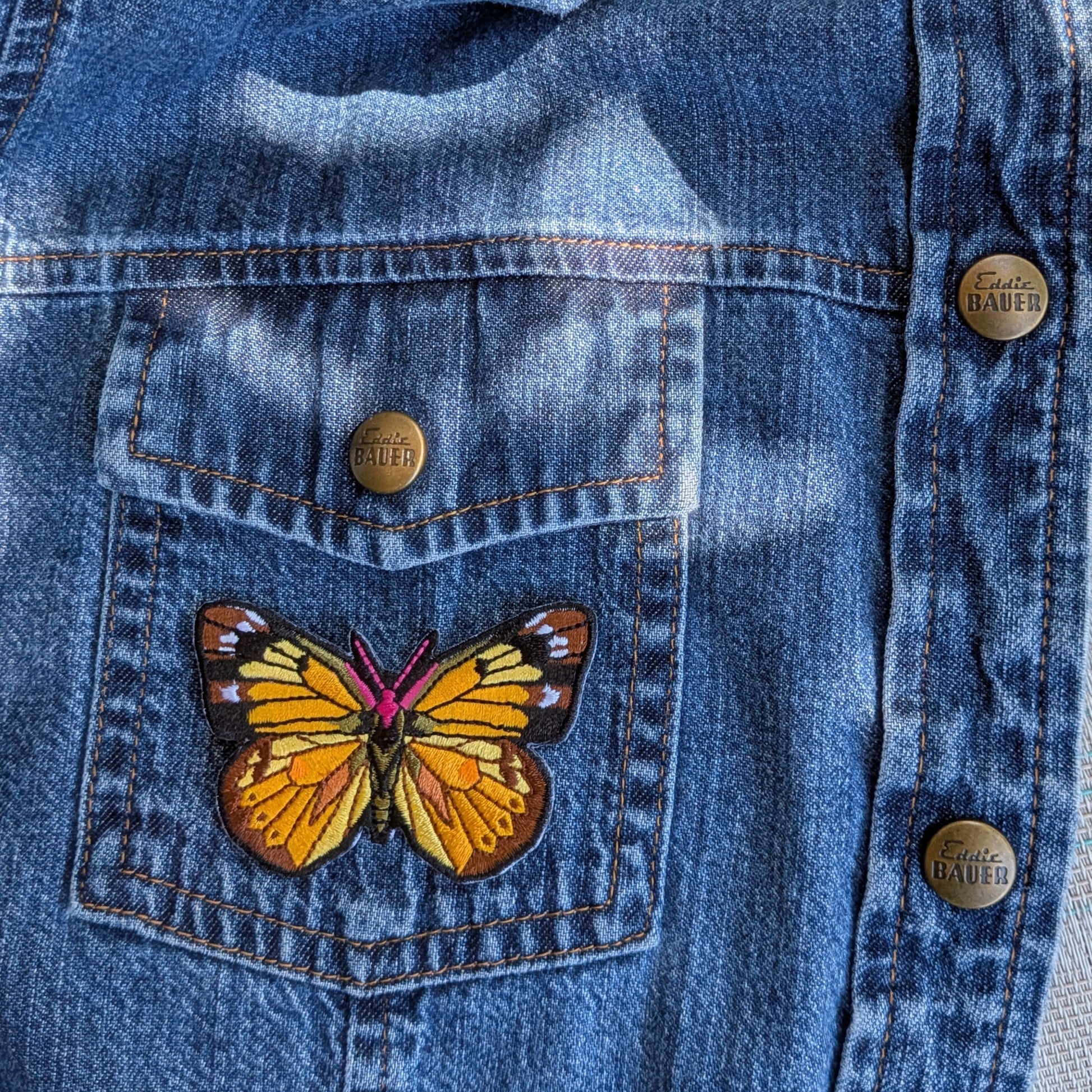 Close-up of a blue denim jacket laid flat outdoors, showing a front pocket with a colorful embroidered patch of a cloused-yellow butterfly. 