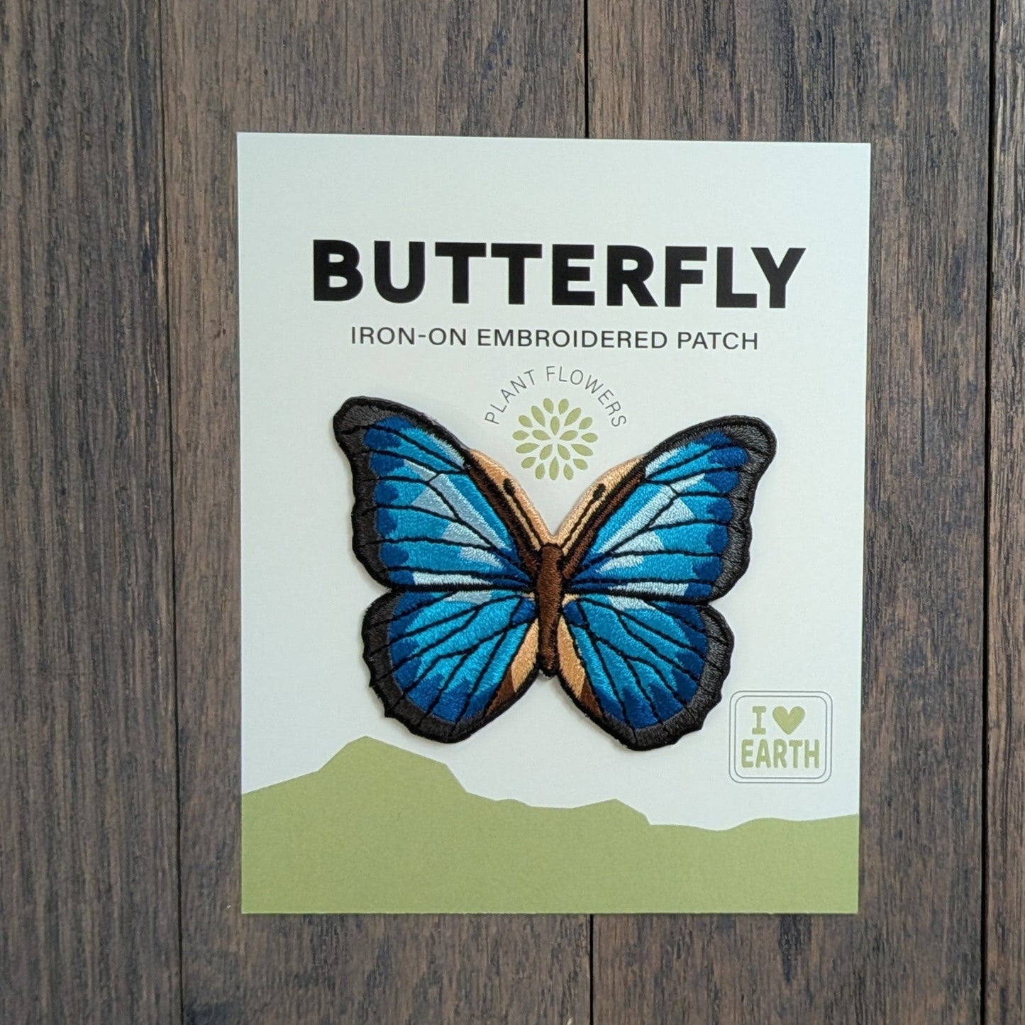 Blue Morpho Butterfly iron-on embroidered patch displayed on a backing card, featuring iridescent blue and black wings with tan accents, on a rustic wood surface.