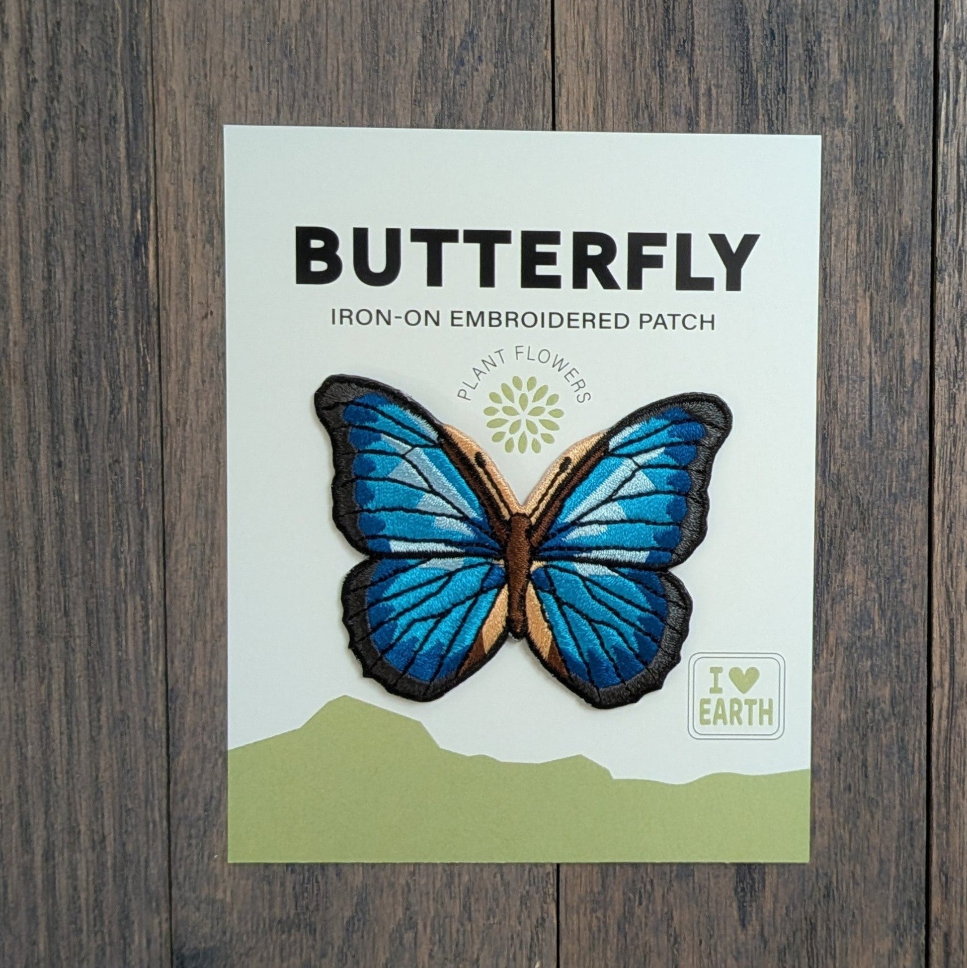 Blue Morpho Butterfly iron-on embroidered patch displayed on a backing card, featuring iridescent blue and black wings with tan accents, on a rustic wood surface.