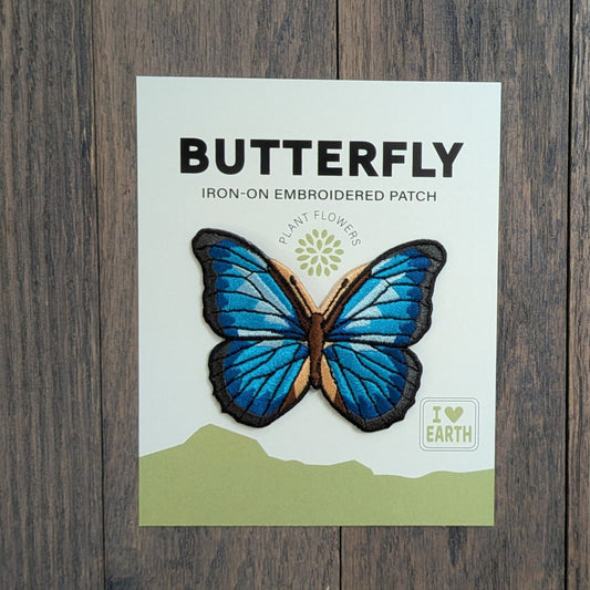 Blue Morpho Butterfly iron-on embroidered patch displayed on a backing card, featuring iridescent blue and black wings with tan accents, on a rustic wood surface.