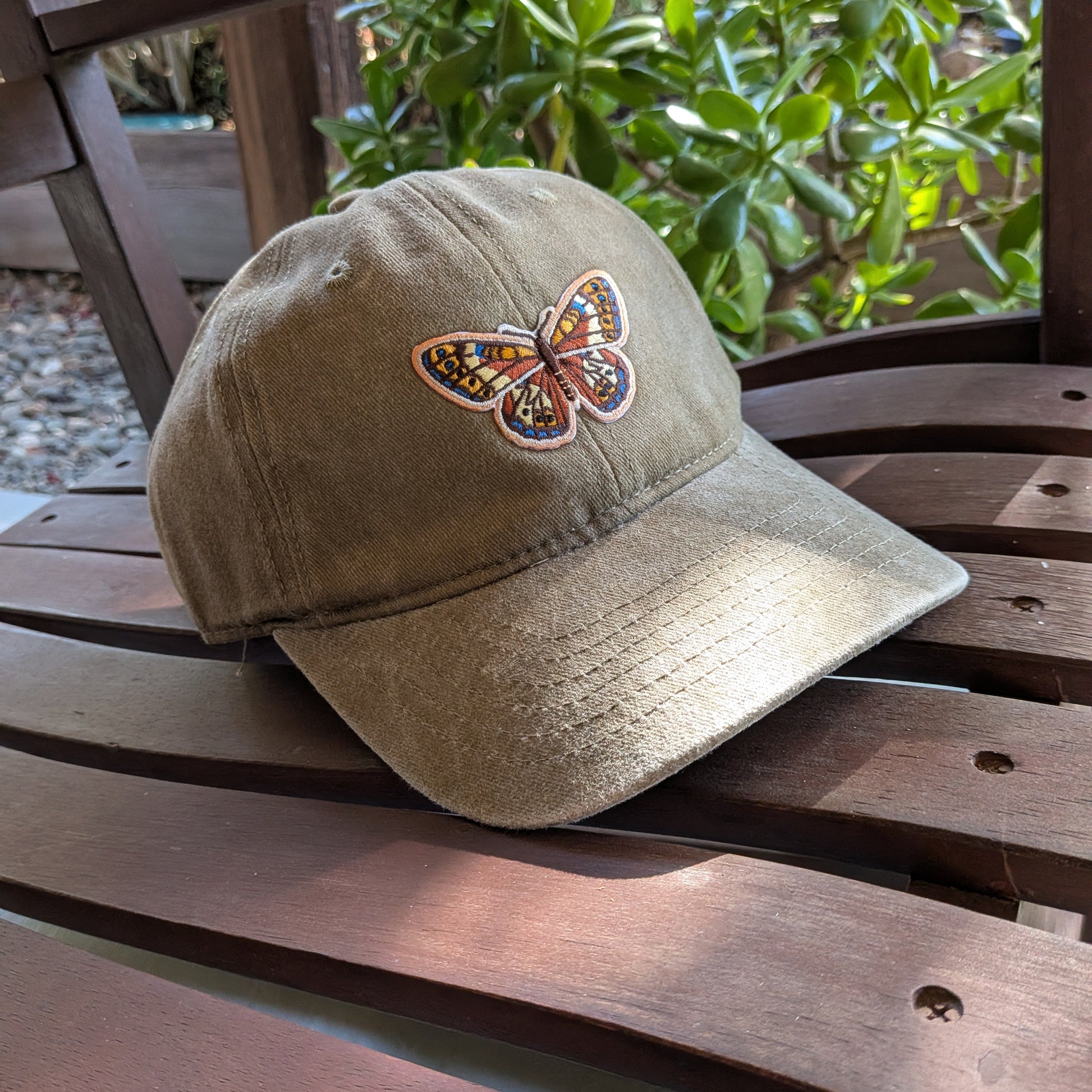 Beige baseball cap with a Metalmark butterfly iron-on embroidered patch on the front panel, styled with wildflowers, lavender, and river stones on a natural wood surface.